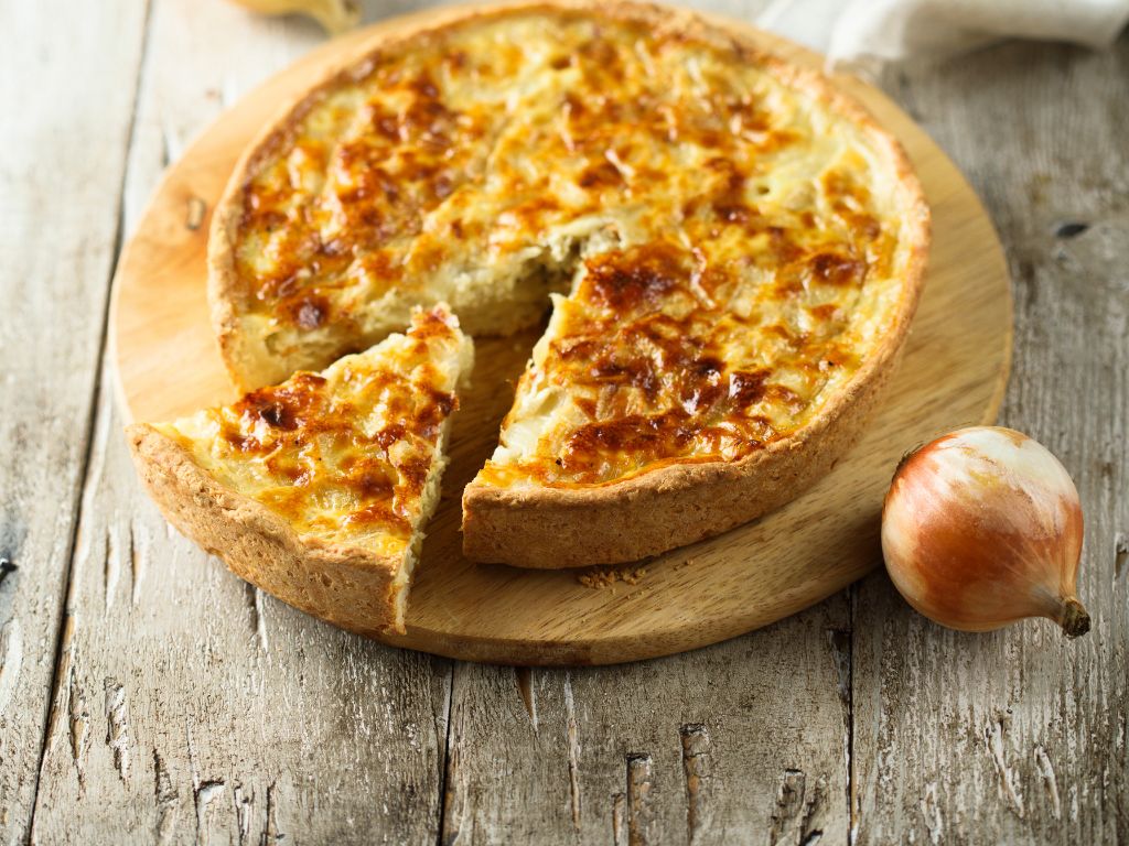 Ein Zwiebelkuchen ist abgebildet: auf einem Tarte-Boden sind Zwiebeln mit Käse überbacken.