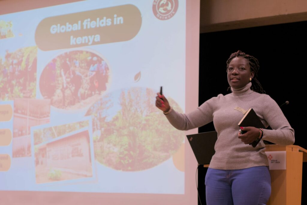 Mercy Ambani steht auf der Bühne und hält einen Vortrag. Im Hintergrund ist eine Präsentation mit der Überschrift "Global Fields in Kenya" zu sehen.