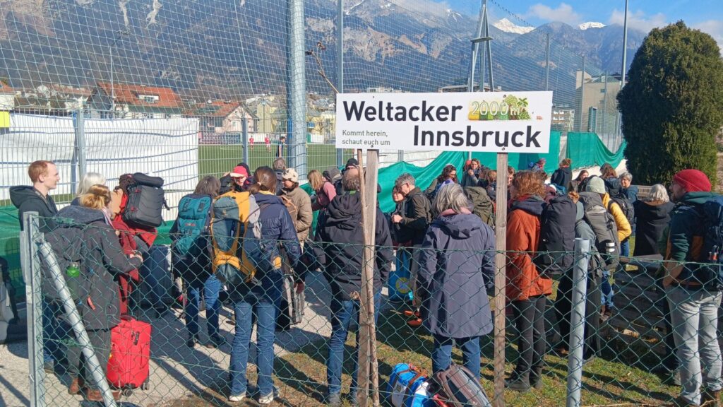 Am Eingang des Weltackers steht ein Schild mit der Aufschrift "Weltacker Innsbruck - Kommt herein, schaut euch um". Drumherum stehen zahlreiche Menschen von Weltäckern mit Gepäck.