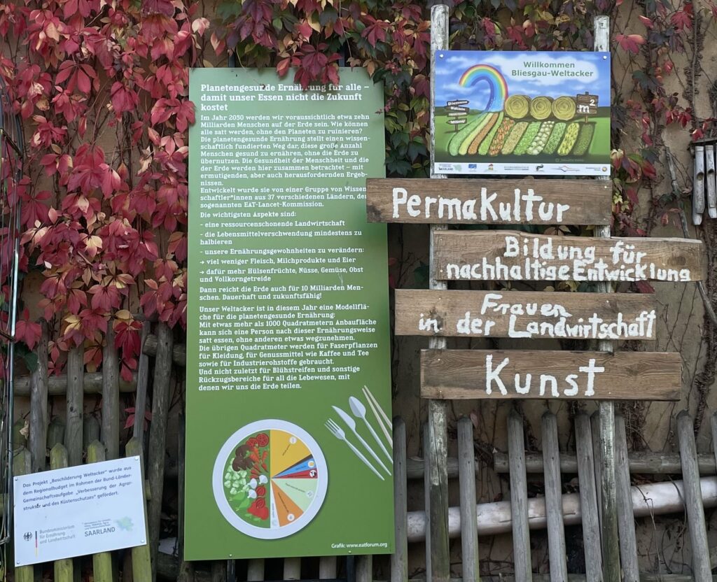 Am Eingang ist ein Schild "Willkommen auf dem Bliesgau-Weltacker" angebracht. Darunter stehen Holzschilder mit "Permakultur", "Frauen in der Landwirtschaft", "Kunst" und "Bildung für nachhaltige Entwicklung". Außerdem steht ein Schild mit einem langen Text zur "Planetengesunden Ernährung".