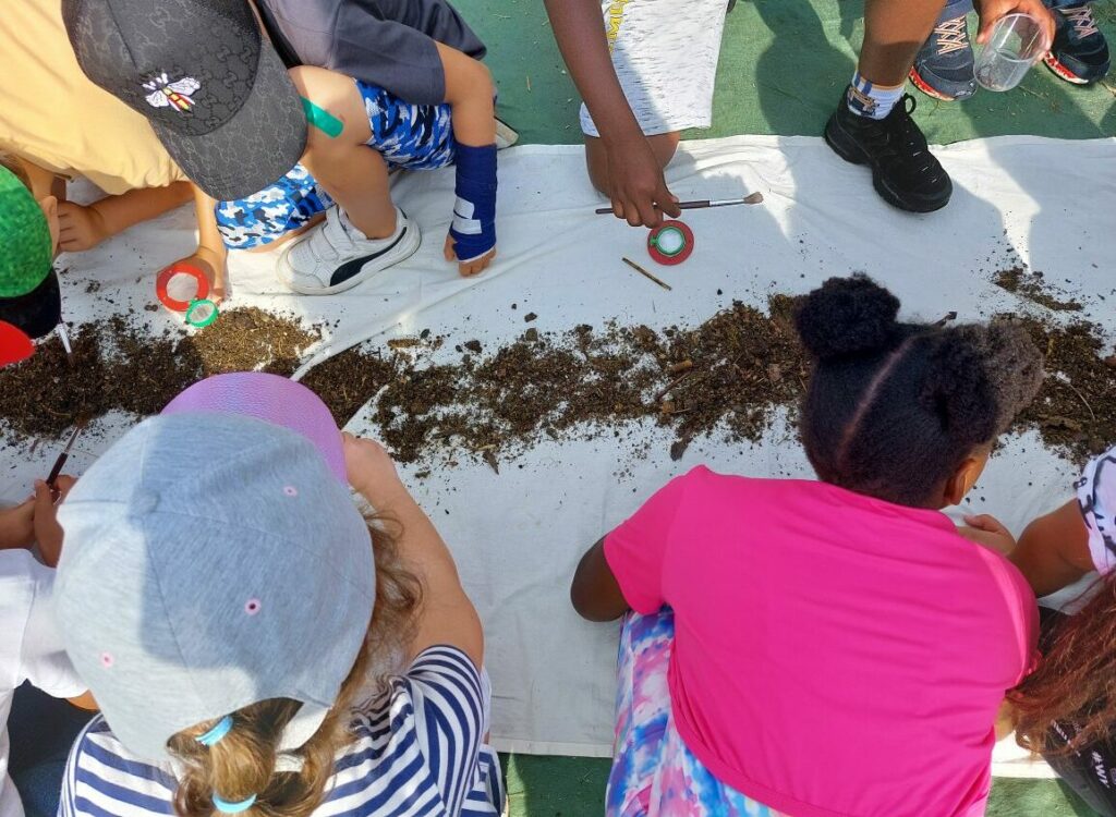 Several children sit on a tarpaulin with brushes in their hands and look at the soil laid out on the tarpaulin.