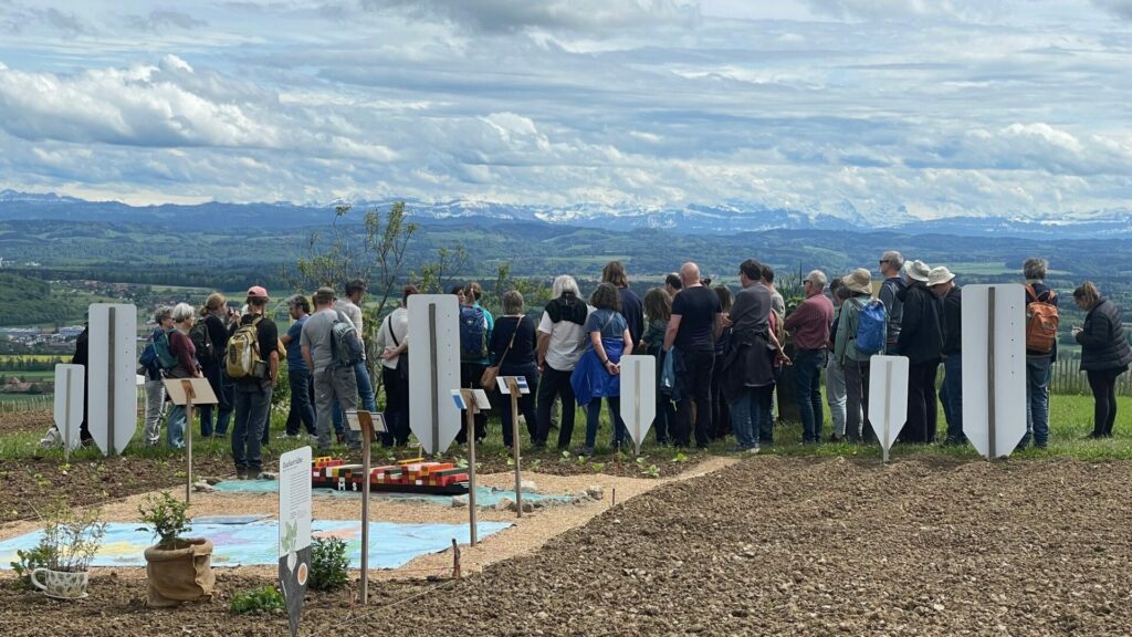 Eine Gruppe lauscht im Frühling einer Ackertour auf dem Weltacker. Im Hintergrund sieht man die Alpen und der wolkenverhangene Himmel.