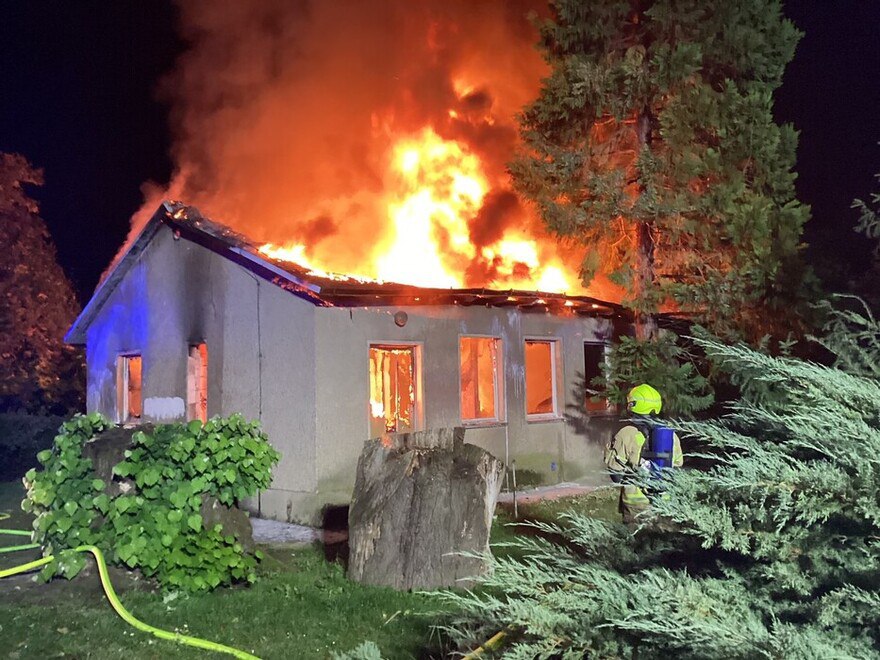 The field hut is completely engulfed in flames at night. A firefighter is standing in front of the house.